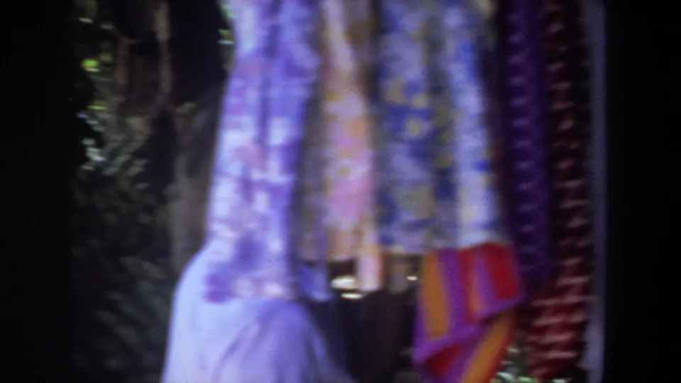 Colorful hanging textiles dominate the frame as the woman in a gray hat remains partly visible at the outdoor Pretoria market.