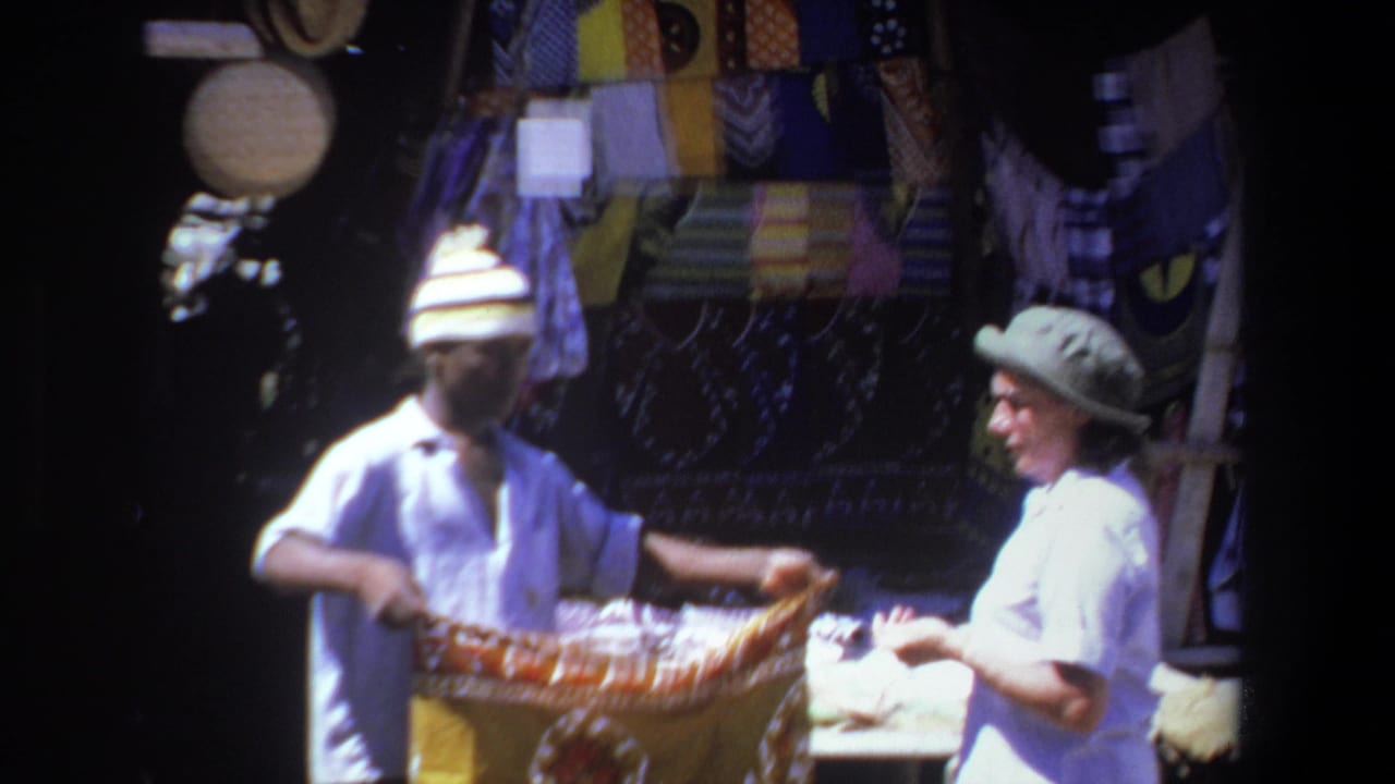 In Pretoria, South Africa, a vendor and customer examine a patterned textile at a shaded market stall in 1969 home-movie footage.