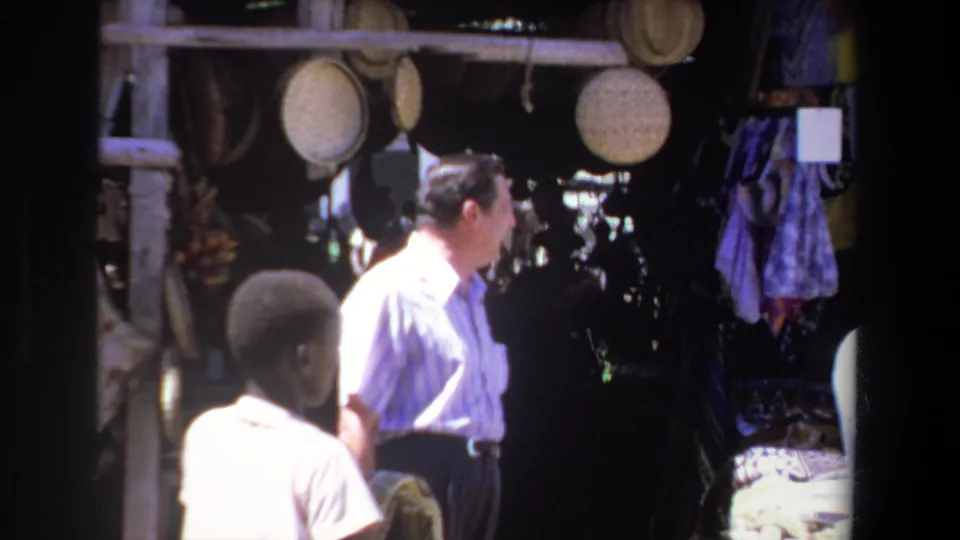 Two people are seen at a Pretoria street market stall with hanging woven baskets and fabric displays in 1969 8mm footage.
