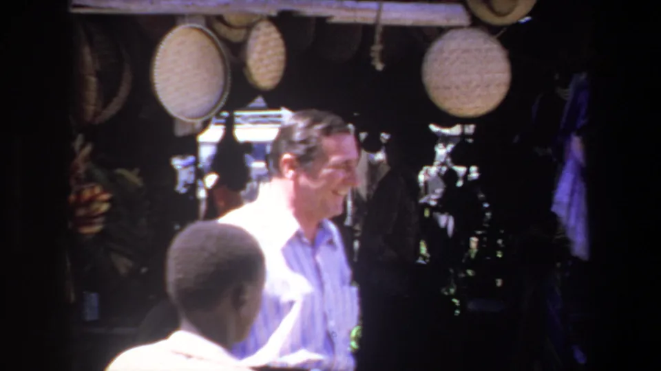 A smiling man in a light shirt stands near a foreground figure beneath woven baskets at a Pretoria market stall, supported by the 1969 metadata.