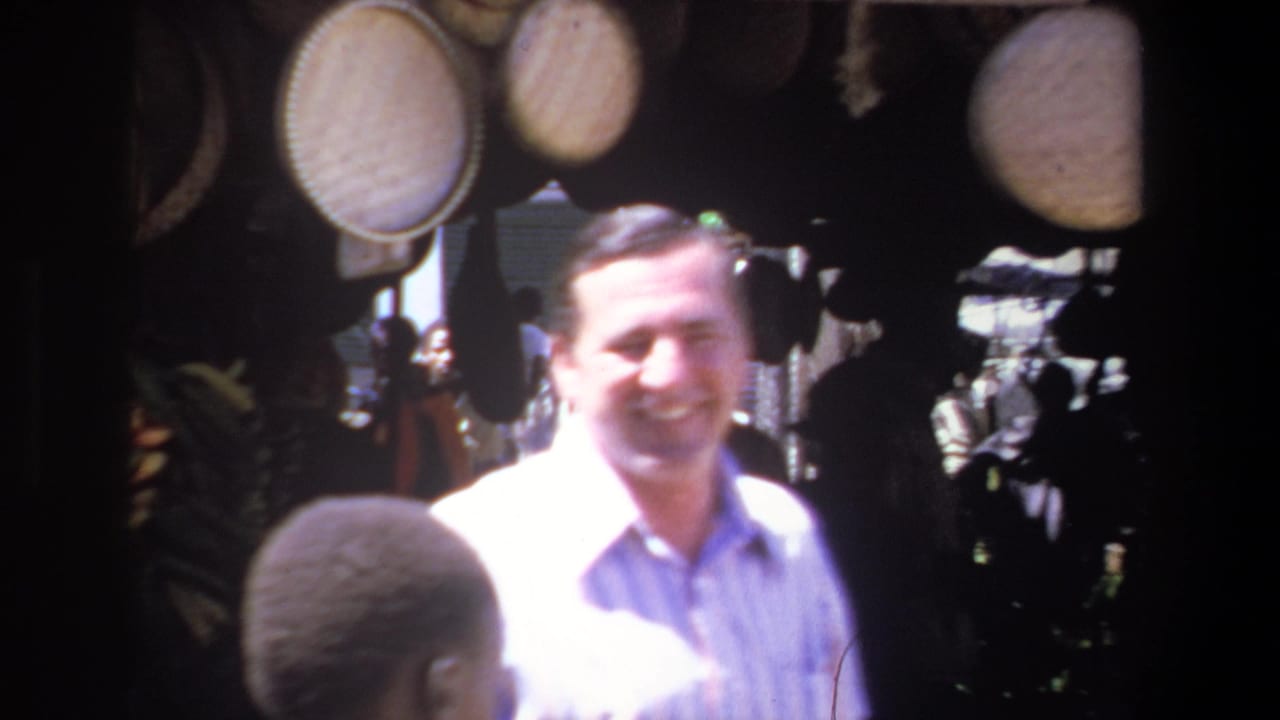 1969 Pretoria home-movie frame showing a smiling man in a striped shirt beside hanging baskets and shaded market goods.