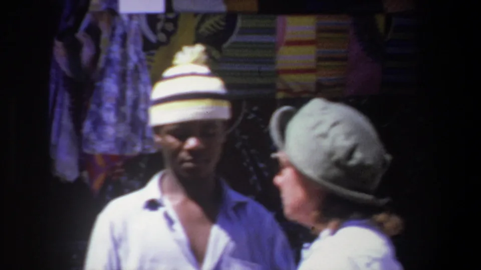 1969 Pretoria market frame with a man in a striped knit hat and a woman in a pale wide-brimmed hat near hanging textiles.