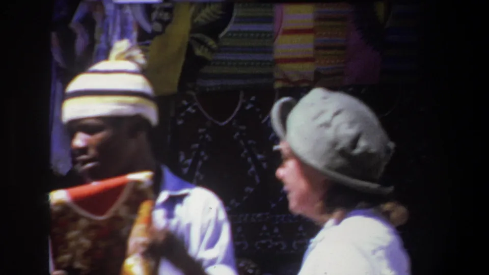1969 Pretoria market frame showing a man holding a folded textile while speaking with a hatted woman near patterned blankets.