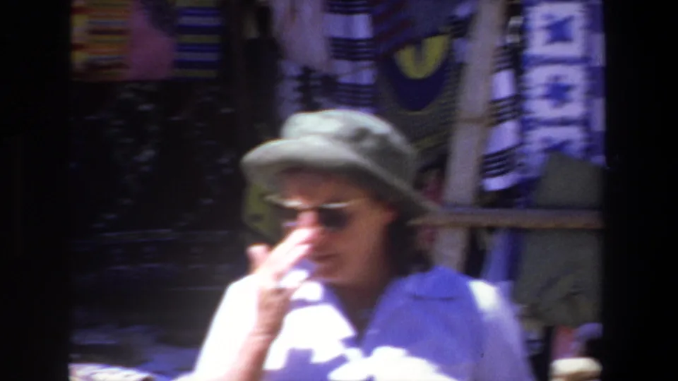 1969 Pretoria home-movie frame of a woman in sunglasses and a wide-brimmed hat standing near a textile display.