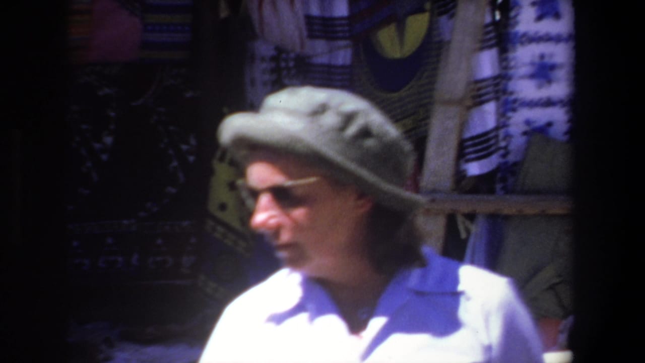 In Pretoria, South Africa, 1969, a woman in a brimmed hat and sunglasses moves past a shaded stall with hanging textiles.