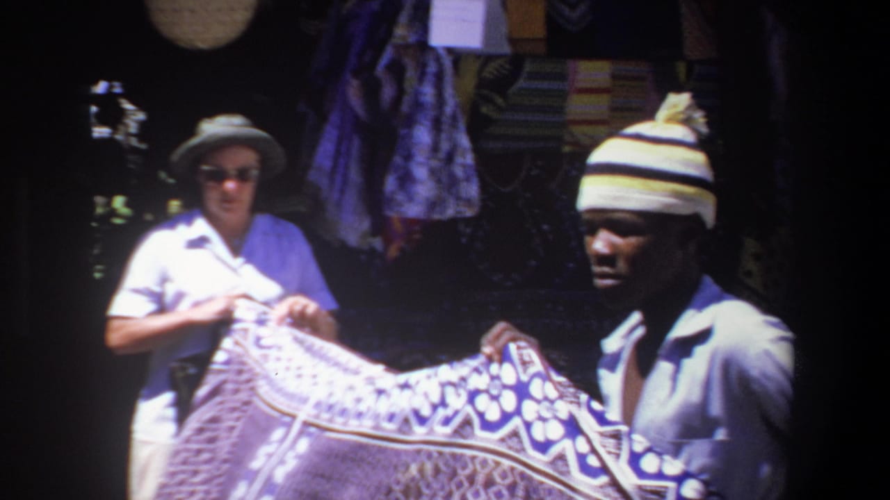 Vendor holds a patterned textile while a woman examines it in a shaded stall.