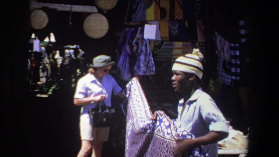 The 1969 Pretoria home-movie view shows a vendor holding a patterned textile while a woman inspects goods nearby.