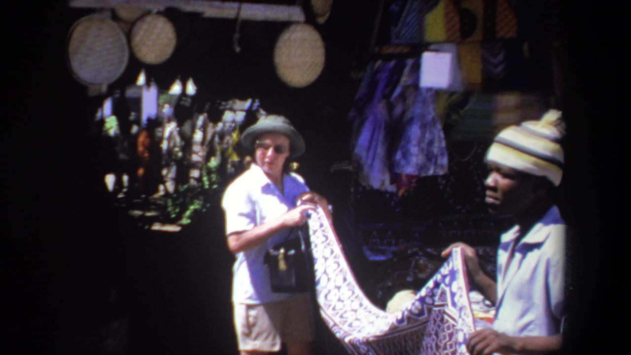 1969 South Africa home-movie frame showing a tourist woman in a sun hat looking at patterned textile with a vendor beside hanging goods.