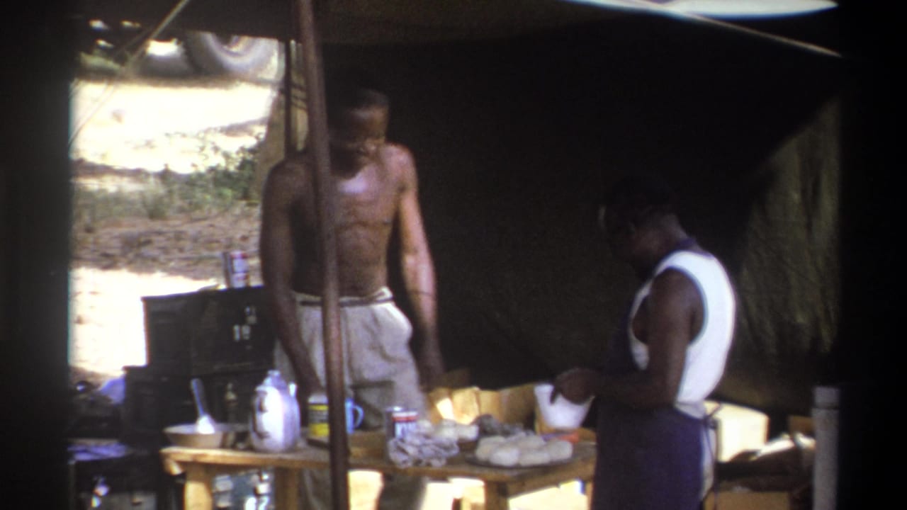 In Cape Town, South Africa, 1969, two adult men work around a food table under a shaded canvas shelter.
