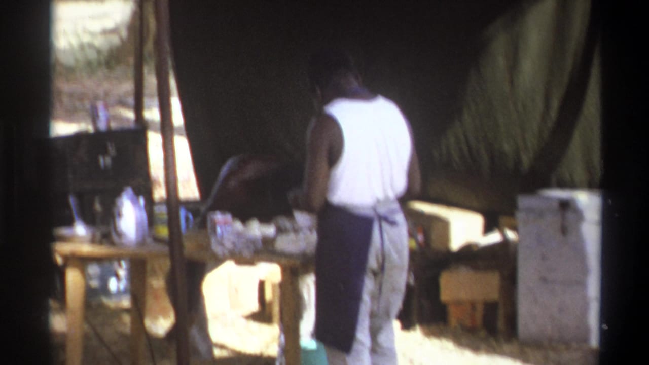 Man in a white tank top stands at a table beneath tenting.