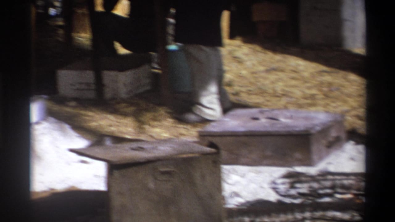 1969 Cape Town home-movie frame showing a rustic camp setup with crates, tent poles and a partly visible adult near outdoor equipment.