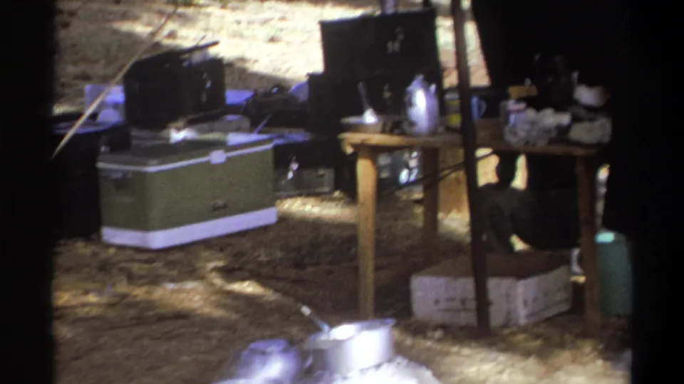 1969 Cape Town home-movie frame showing a green cooler, small table, metal cookware and stacked supplies inside a rustic camp shelter.
