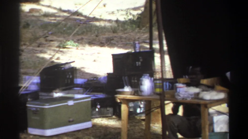 1969 Cape Town camp frame looking through shade at a green cooler, table, kettle, boxes and cookware in a temporary outdoor setup.
