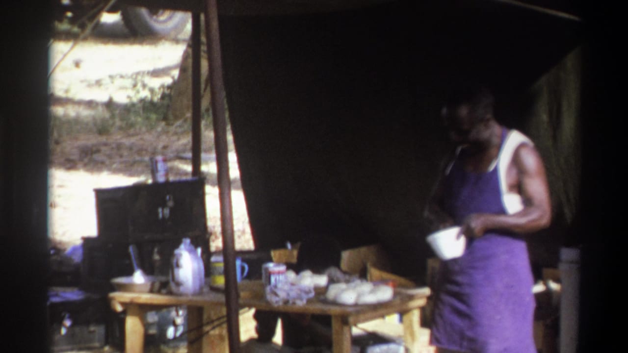 1969 Cape Town home-movie frame of a man in a purple apron holding a white bowl beside a camp table inside a tent.