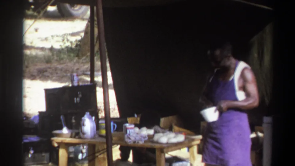 1969 Cape Town home-movie frame of a man in a purple apron holding a white bowl beside a camp table inside a tent.