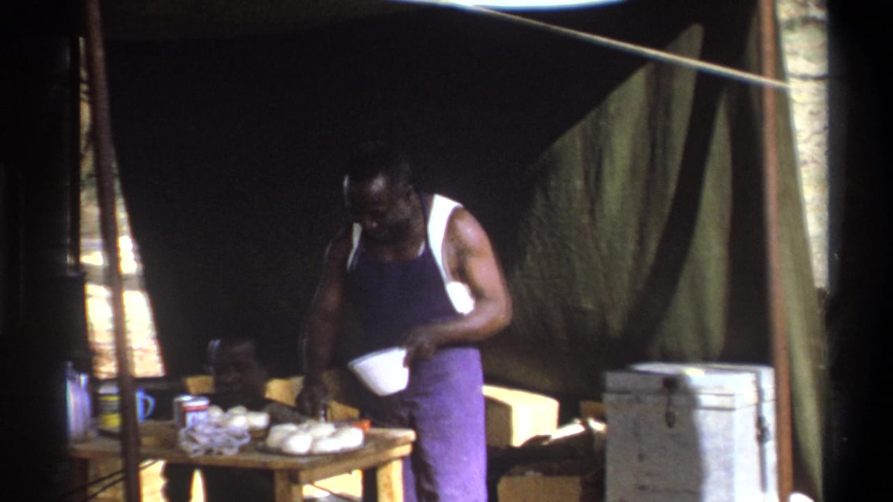 Standing man works with a bowl at a tent table with food and supplies, with a seated man behind him.