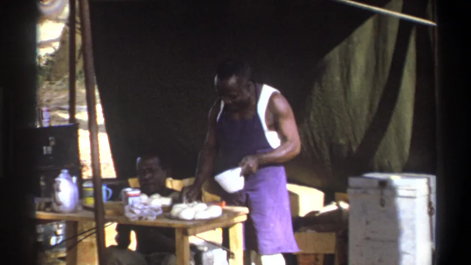 Cape Town 1969 home-movie still of informal meal preparation inside a shaded tent, with camp supplies arranged around a wooden table.
