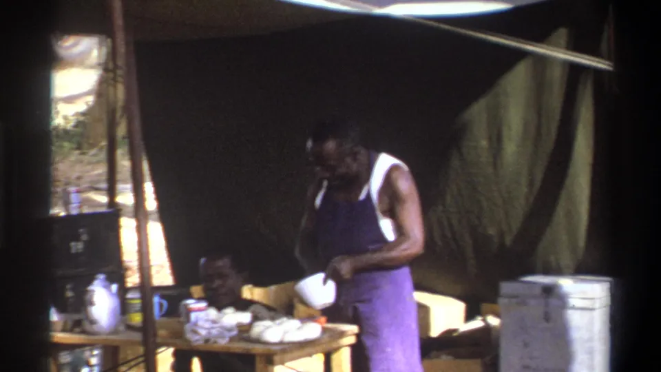 1969 Cape Town 8mm frame of a man cooking or serving food in a tented camp area with a cooler and table supplies nearby.