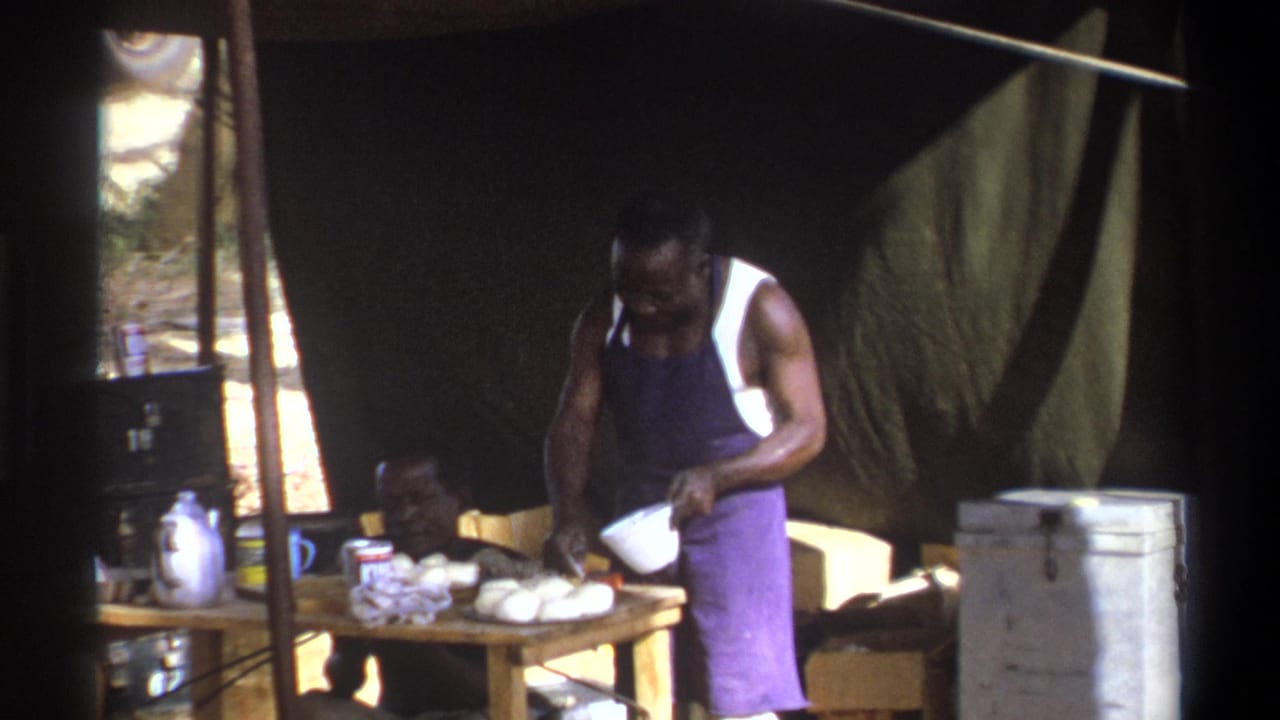 Standing man pours or handles a white bowl at a table while another man sits nearby.