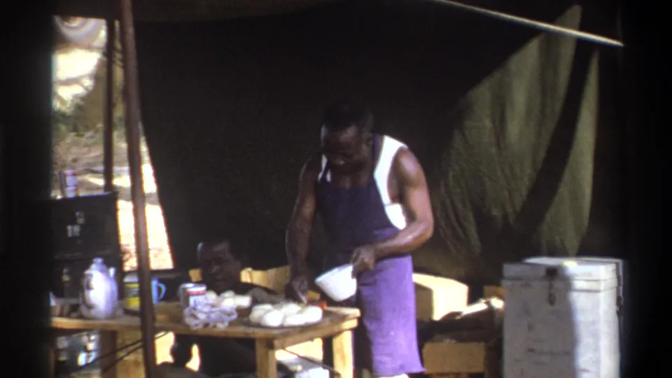 1969 Cape Town home-movie still of a man in a white tank top and purple apron working with a bowl at an outdoor tent table.