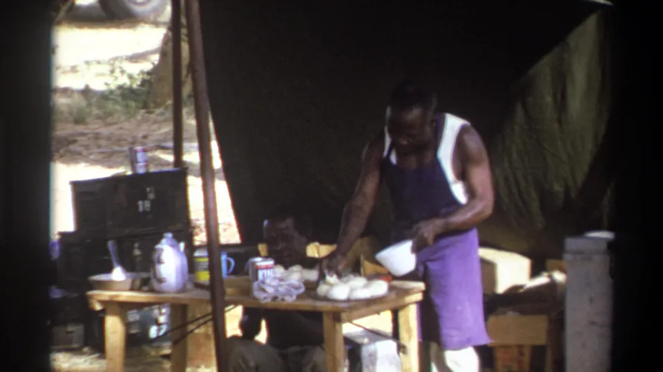 1969 Cape Town home-movie frame of two men at a makeshift outdoor kitchen under a large canvas tent.