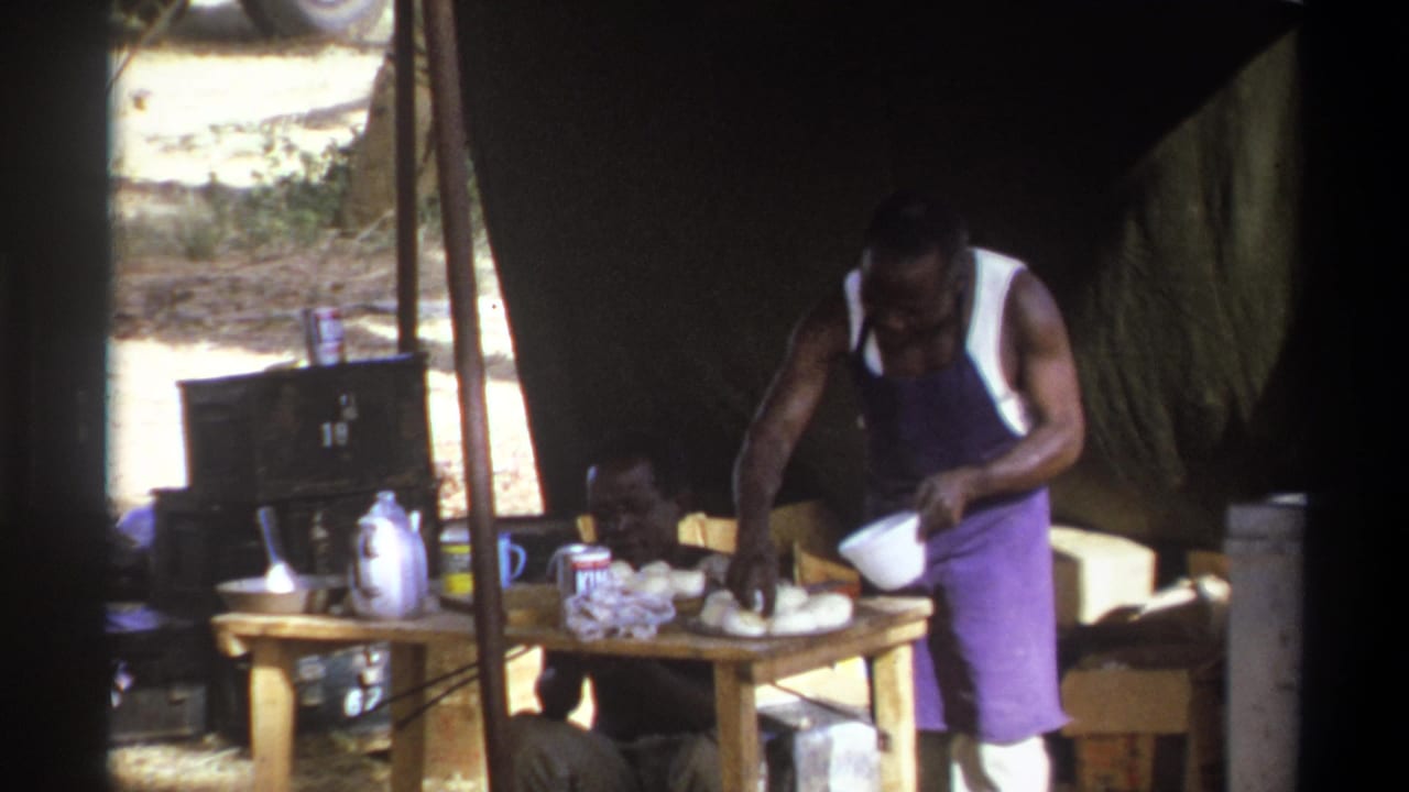 1969 home-movie frame from South Africa showing a person at a camp table under shade, with containers and luggage around the work area.