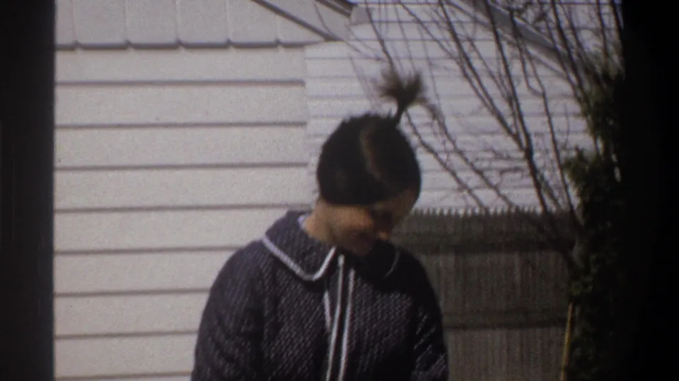 1969 home-movie frame showing a woman in a dark blue robe near a snow-lined driveway outside a white-sided house.