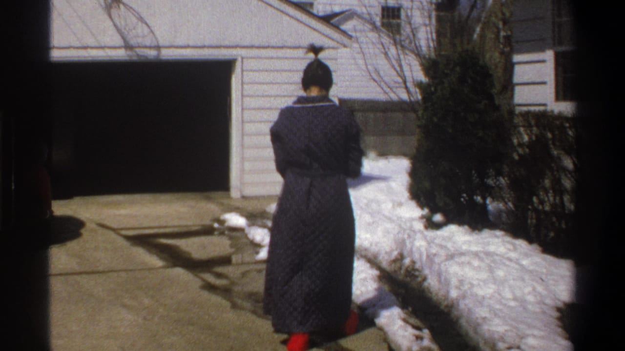 1969 home-movie frame of the woman from behind on a driveway with snowbanks, shrubs, and an open garage nearby.