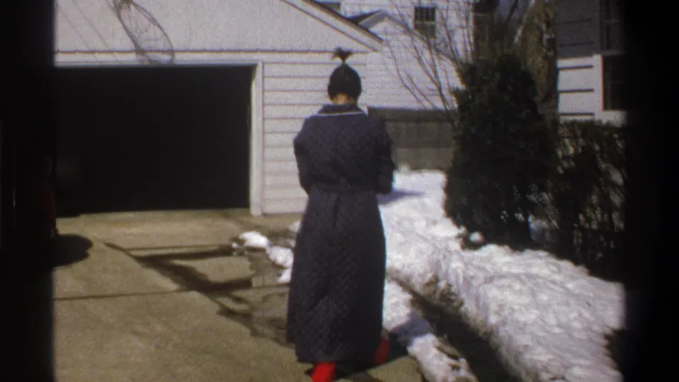1969 home-movie frame of the woman from behind on a driveway with snowbanks, shrubs, and an open garage nearby.