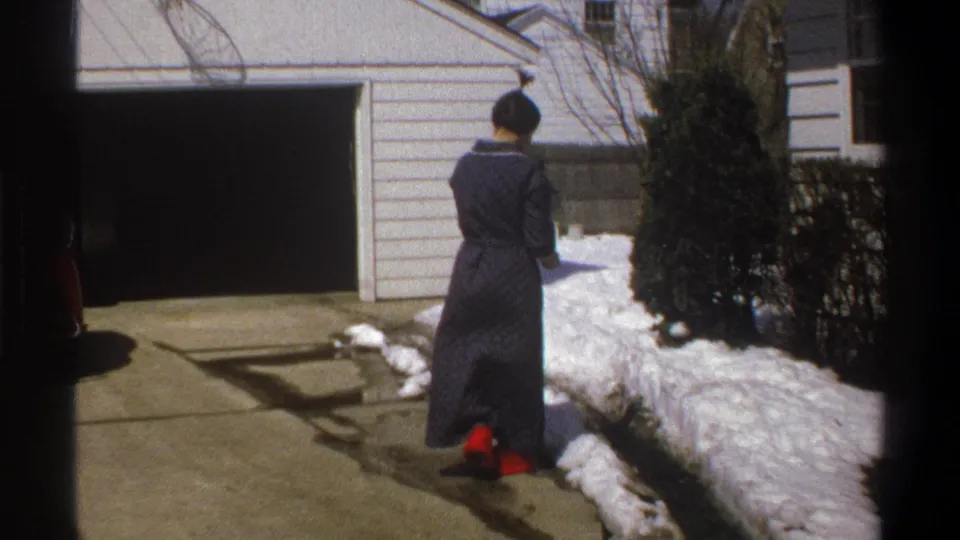 1969 home-movie frame showing the robed woman paused from behind beside a snow-covered edge of the driveway.