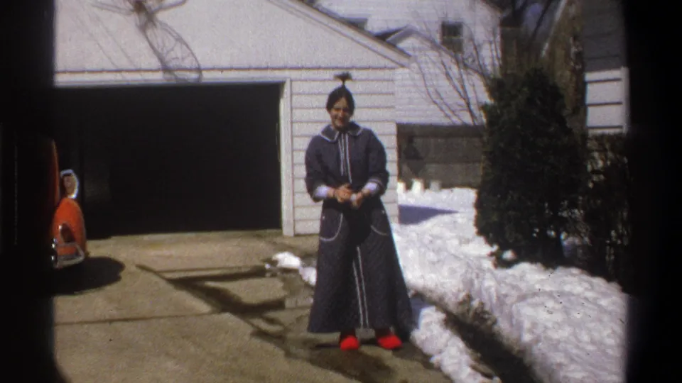 1969 Cape Town home-movie frame of a woman in a dark robe facing the camera while holding a snowball in the driveway.