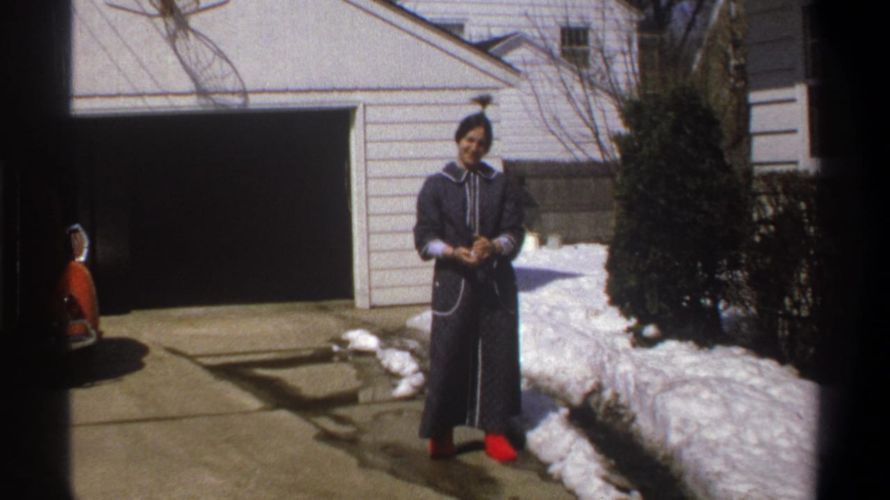 Woman in a dark robe and red shoes stands in a snowy driveway near a white garage.