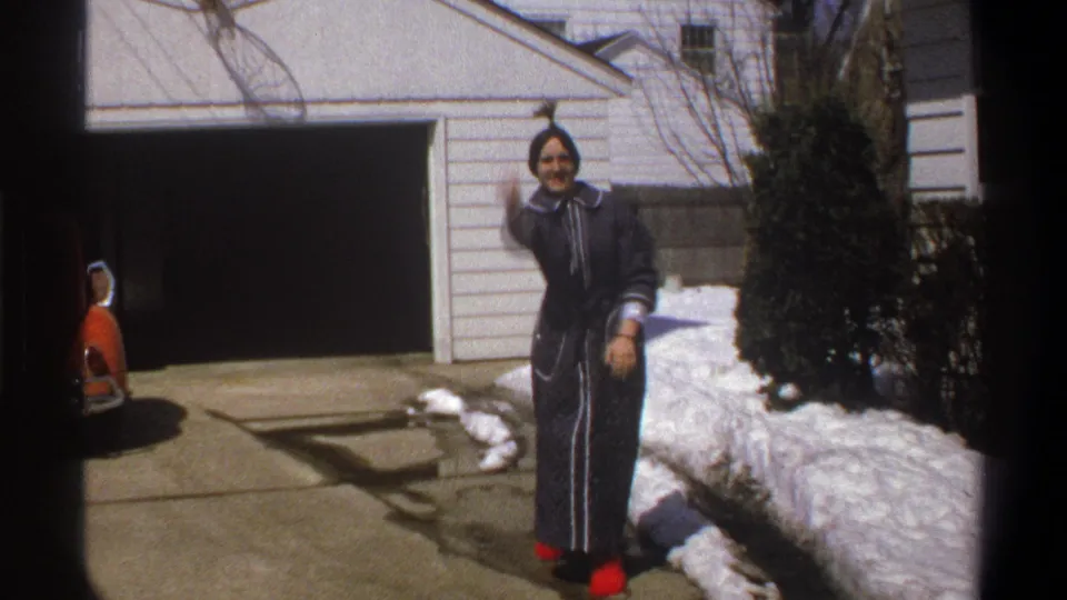 1969 Cape Town home-movie frame showing the woman smiling and raising an arm in the snowy driveway.