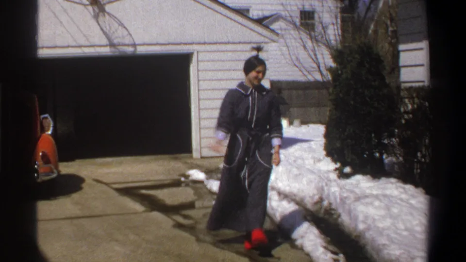 1969 Cape Town frame of the woman moving across a snowy residential driveway in a dark robe and red shoes.