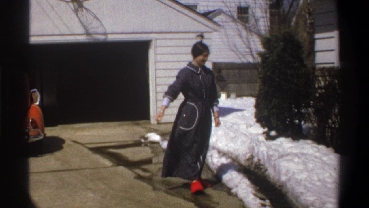 In Cape Town 1969 home-movie footage, a young woman in a dark robe and red shoes stands beside melting snow near a garage.