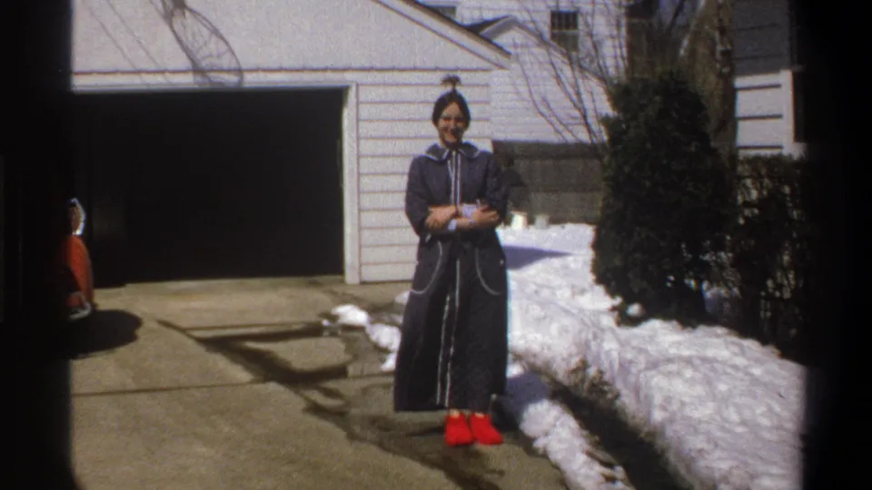 The subject smiles toward the camera in Cape Town 1969 footage, standing by snow on a driveway in red shoes and a dark robe.