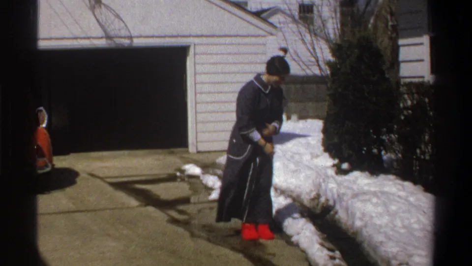 In the 1969 Cape Town home-movie scene, the subject bends slightly and looks down while handling snow beside the driveway.