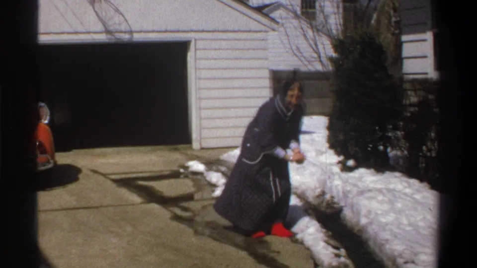 The subject crouches beside melting snow in Cape Town 1969 footage, wearing a dark robe with white trim and red shoes.