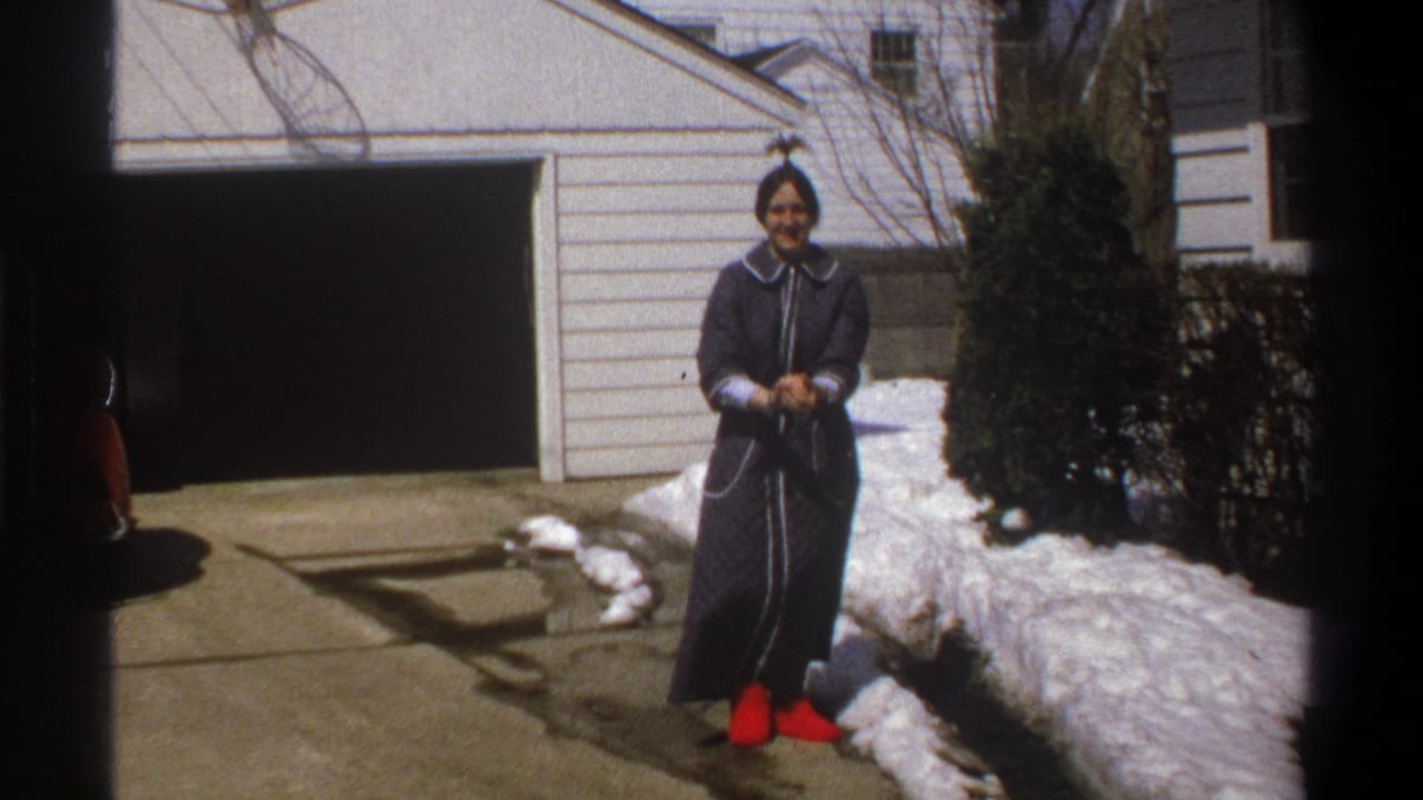 In 1969 Cape Town home-movie footage, a woman in a dark coat and red shoes faces the camera on a snowy driveway by a white garage.