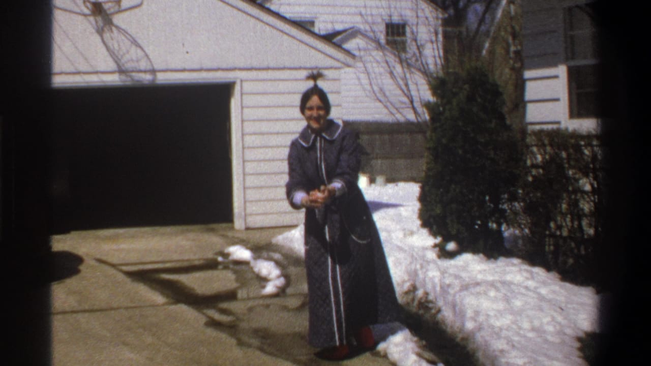Woman smiles while holding a snowball in front of a garage and snow-covered driveway edge.