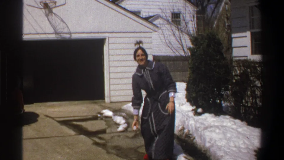 The woman leans into the playful trick on the snowy driveway, with red shoes, garage siding, and a basketball hoop visible behind her.