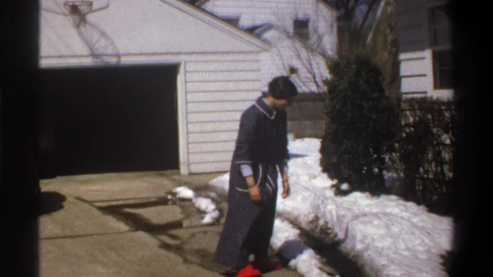 At the end of the trick, the woman looks down near the snowbank beside the driveway and white garage in the 1969 home-movie scene.