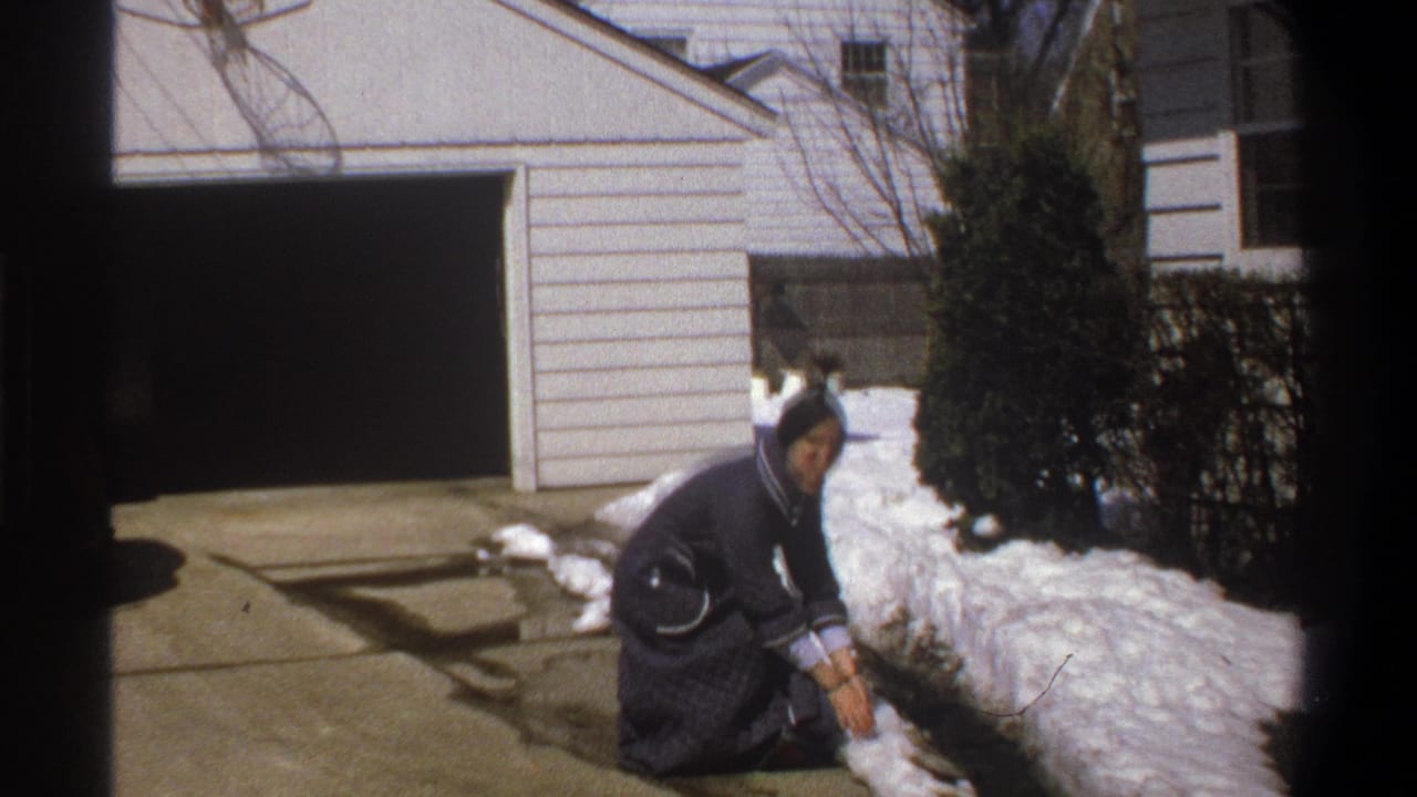 In 1969 Cape Town home-movie footage, a woman in a dark blue dress crouches by snow beside a driveway and garage.