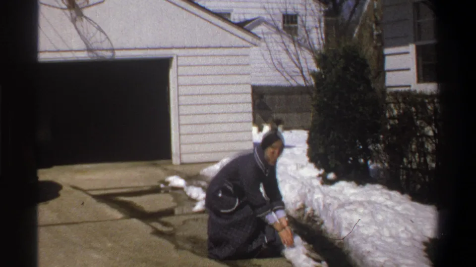 In 1969 Cape Town home-movie footage, a woman in a dark blue dress crouches by snow beside a driveway and garage.