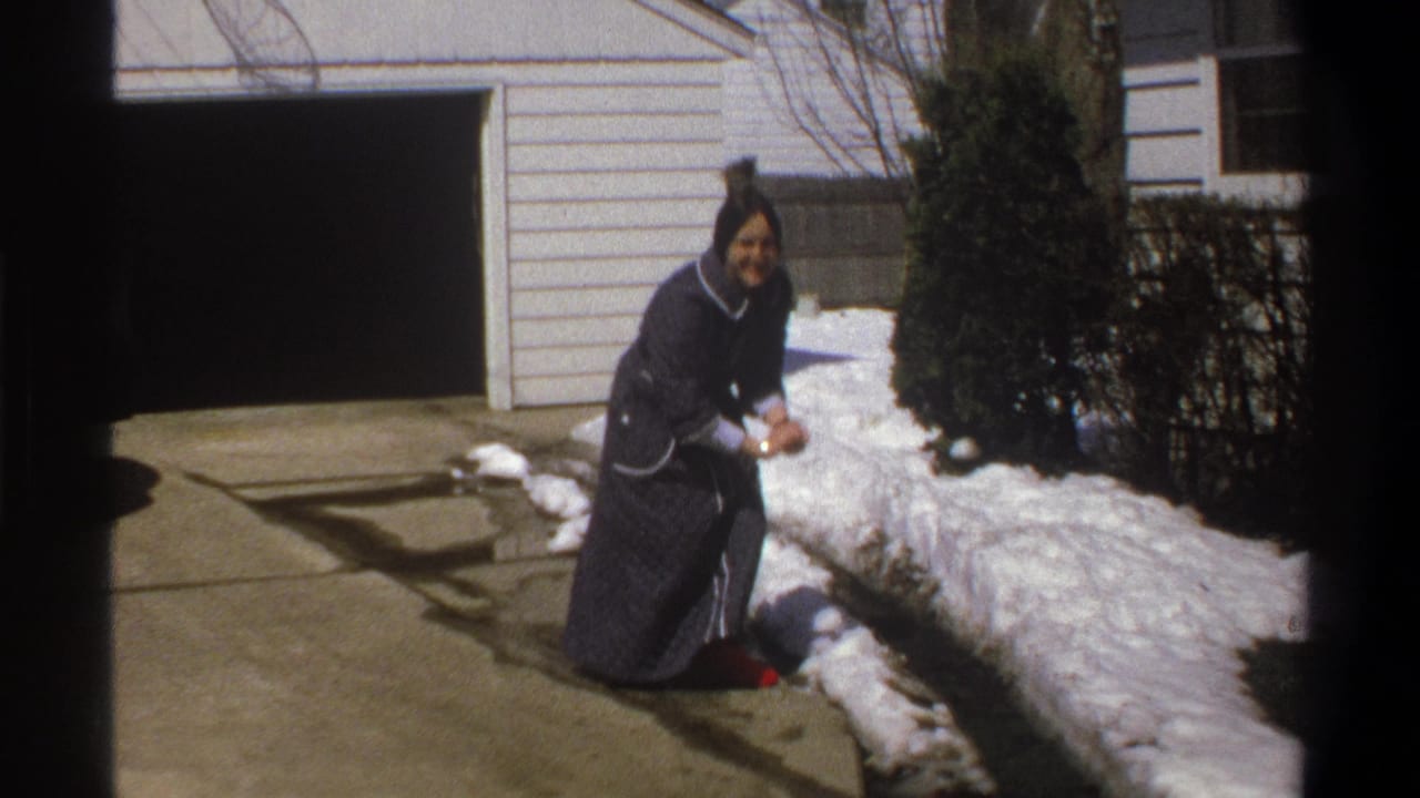 Woman kneels by snow in front of a white-sided house and garage.
