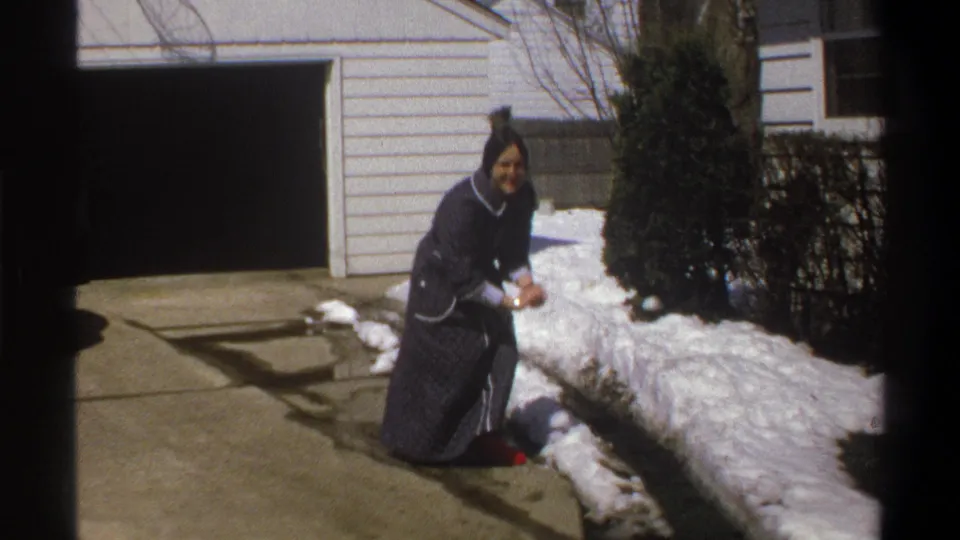 The woman kneels near piled snow outside a white-sided house, holding snow as she faces the camera.