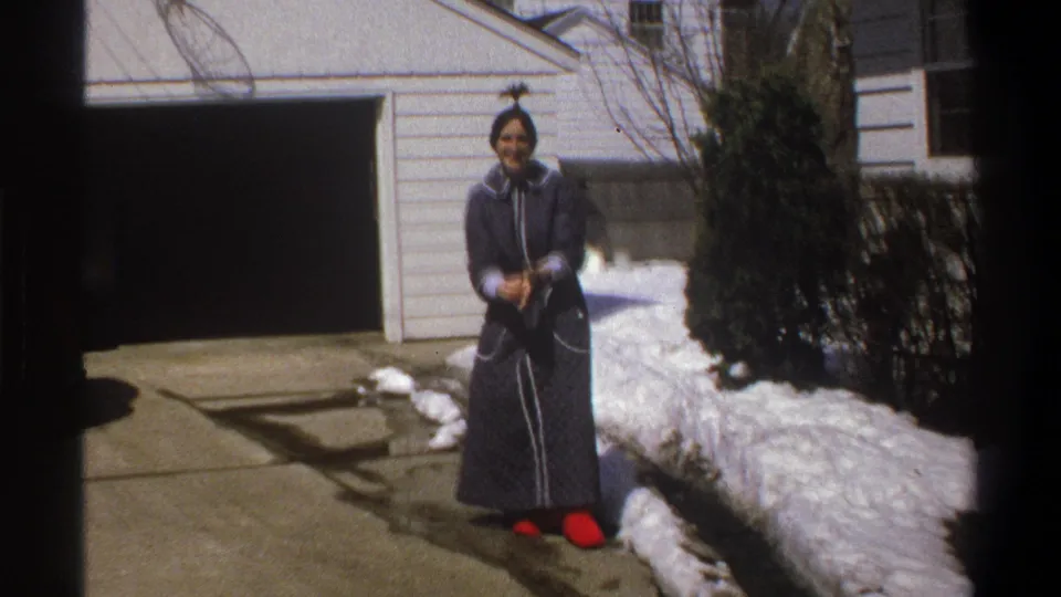 The woman stands from the snowbank in a dark quilted dress and bright red shoes, smiling toward the home-movie camera.