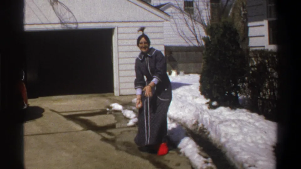 A smiling woman in a dark blue dress and red shoes stands beside snow in a Cape Town driveway, framed by shrubs and a garage.