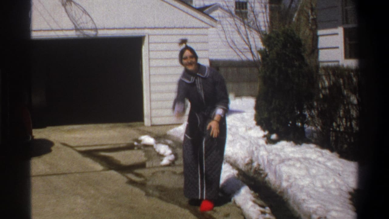 Woman in a dark robe stands by a snowy driveway near a house.