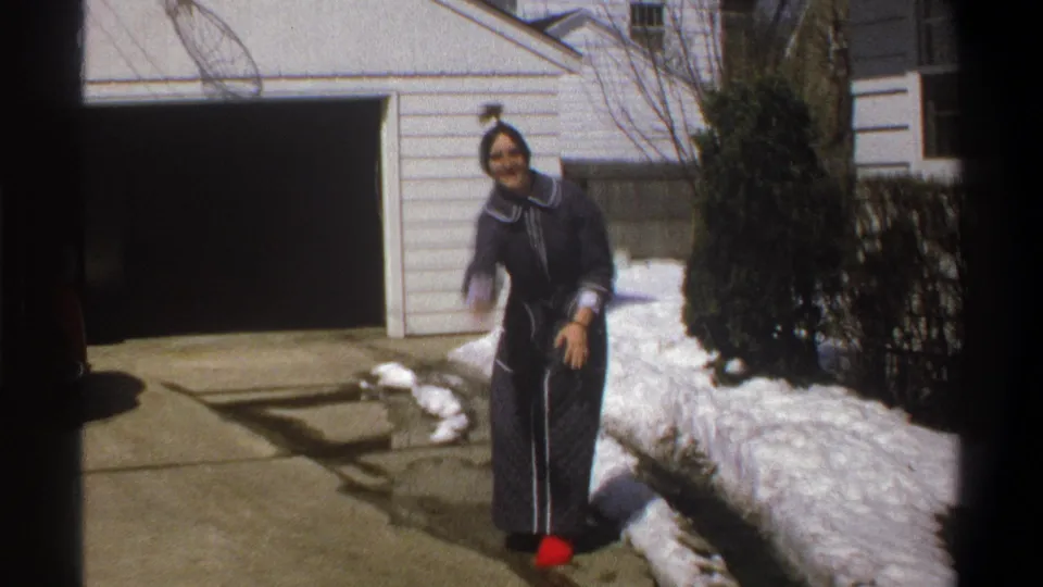 In 1969 Cape Town home-movie footage, a woman in a dark robe and red slippers stands by snow along a residential driveway.